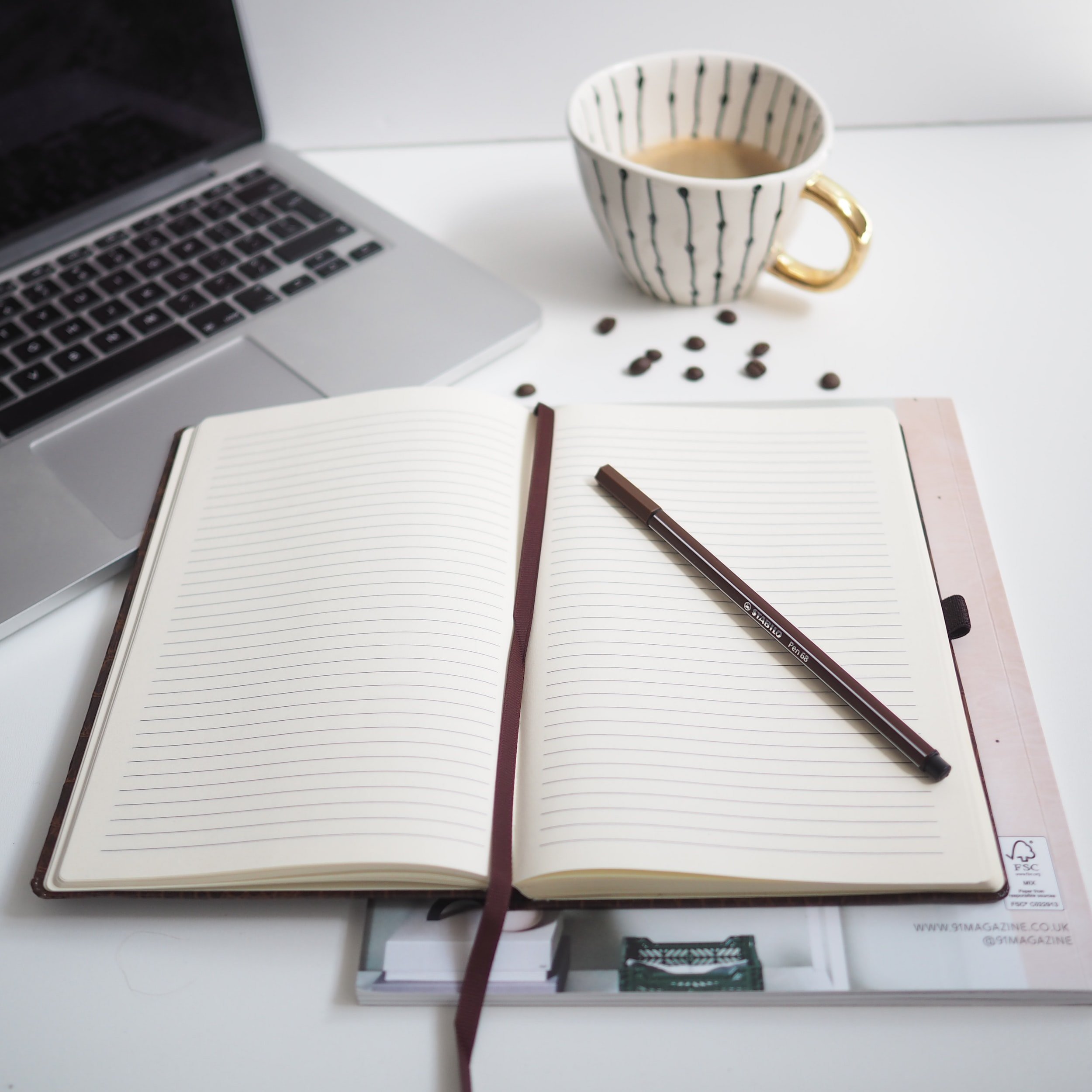 A notebook and pen, coffee mug, and laptop sitting on a white desk